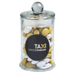 A clear glass apothecary jar filled with yellow and white chocolate beans, featuring a round black logo.