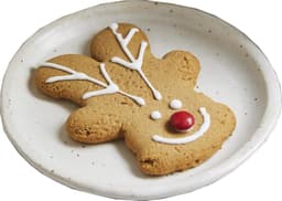 A gingerbread reindeer cookie in a light brown colour, decorated with white icing and a red candy on a white plate.