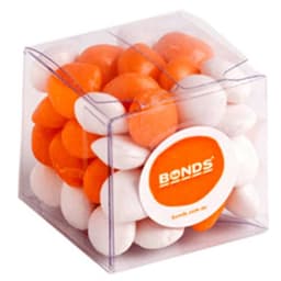 A cube-shaped container filled with orange and white chewy fruit candies. The container has a branding logo on it.