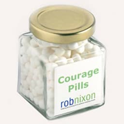 A square glass jar with a gold lid, filled with white mints. The jar features a label with a logo.