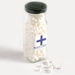 A tall glass jar with a black lid filled with white mints, partially spilled onto a light background.