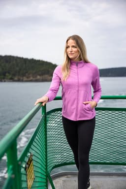 A woman stands on a boat wearing a pink full zip jacket and black leggings, with water and trees in the background.