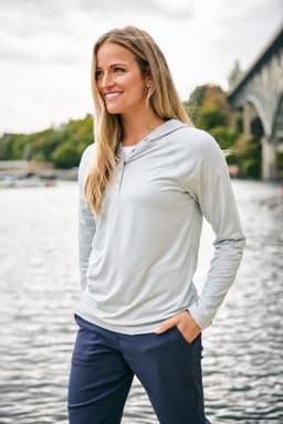 A hooded shirt in light grey, featuring long sleeves and a relaxed fit, displayed by a model near water.