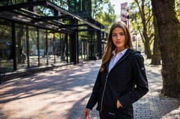 A woman stands outside wearing a black softshell jacket. The jacket features a hood and has a logo.