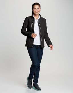 A woman wearing a black softshell jacket over a white top and jeans, featuring a logo on the jacket.