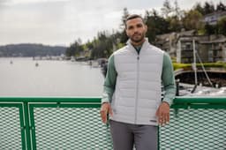 A man wearing a white eco hybrid vest over a green top, standing on a green railing with a waterfront background.