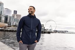 A man wearing a navy blue eco-friendly rain jacket stands by the water with a city skyline in the background.