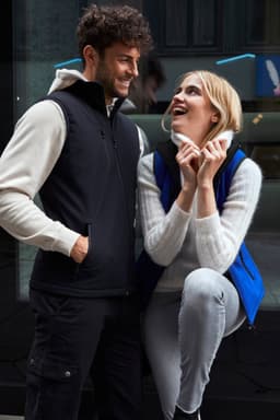A black softshell vest is worn by a man, while a woman smiles in a blue softshell vest, both against a modern backdrop.