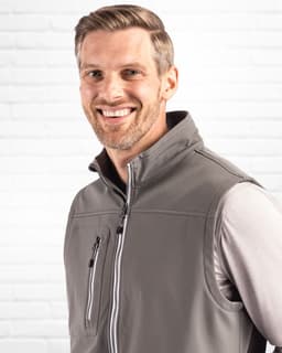 A grey men's softshell vest with a high collar and zip pockets, modelled by a smiling man against a white brick background.