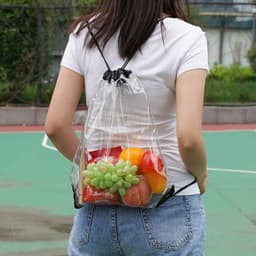 A transparent drawstring backpack filled with assorted fruits, held by a person wearing a white shirt and denim shorts.