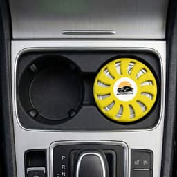 A round, yellow air freshener is placed in a car cup holder, featuring a logo and a vented design.