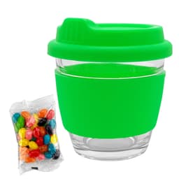 A reusable coffee cup with a clear glass body and a bright green silicone lid and sleeve, alongside a packet of colourful jelly beans.