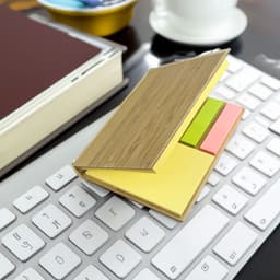 A bamboo sticky note holder with yellow, green, and pink sticky notes, placed on a white keyboard beside a book and a cup.