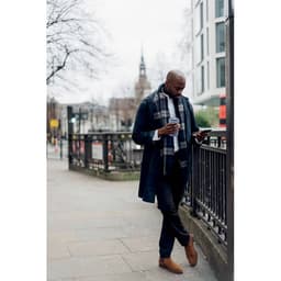 A man in a long navy coat and grey scarf holds a coffee cup while looking at his phone on a city street.