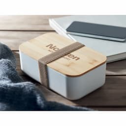 A stylish white and bamboo lunch box with a fabric strap, placed on a wooden surface beside a grey knit and phone.