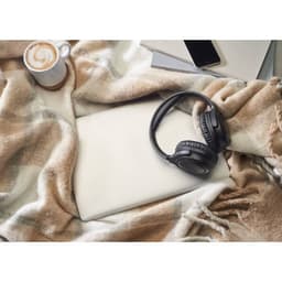 A flat lay featuring a laptop, black headphones, a coffee cup, and a patterned blanket in neutral colours.