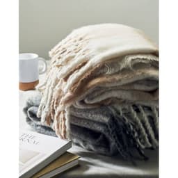 A stack of soft, textured blankets in shades of cream, beige, and grey beside a white mug and a book.