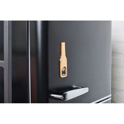 A bottle opener shaped like a drink bottle attached to a black fridge, made from wood. It has a round cutout and a simple design.