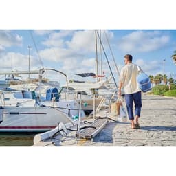 A man carries a blue and white bag along a marina, surrounded by sailboats and a sunny sky.