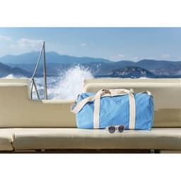 A blue duffel bag with tan straps rests on a sunlit boat seat, beside a pair of sunglasses, against a scenic backdrop.
