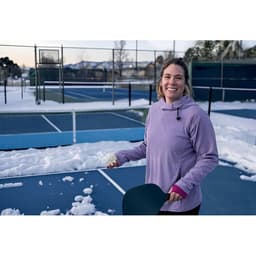 A woman in a lavender hoodie stands on a snowy tennis court, holding a snow shovel and smiling.