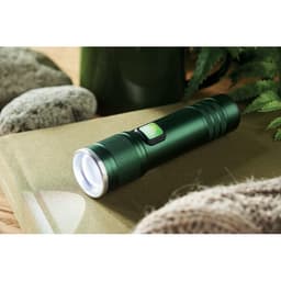 A green flashlight featuring a textured grip and a clear lens, resting on a natural surface with stones.