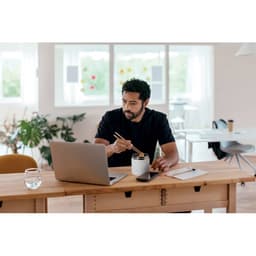 A man with dark hair uses chopsticks while sitting at a wooden table with a laptop, a glass of water, and a notepad.