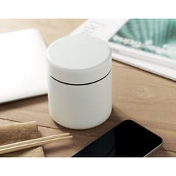 A white container with a lid sits on a wooden table beside a phone and chopsticks, with papers in the background.