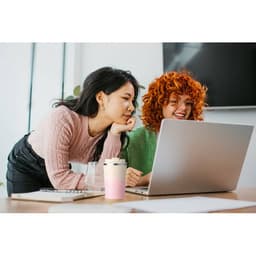 Two women leaning over a laptop, engaged in conversation. One has long dark hair and the other has short curly red hair.