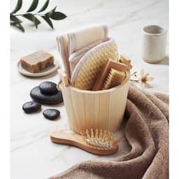 A wooden bucket filled with natural sponges, a brush, towels, soap, stones, and a towel draped beside it.