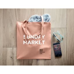 A tote bag in peach colour, with grey items inside, alongside sunglasses and a smartphone. The bag features a logo.