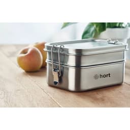 A stainless steel lunchbox with a clasp and branding, placed on a wooden surface beside two apples.