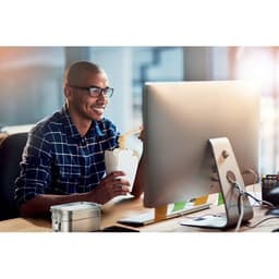 A drink bottle in a silver colour next to a man enjoying a takeaway meal while working at a desk with a computer.