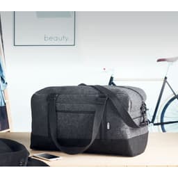 A grey fabric bag with a black base, featuring a shoulder strap and zippers, sits on a table near a bicycle.