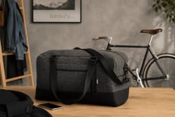 A grey and black duffle bag with sturdy handles, positioned on a wooden table beside a bike and a jacket.