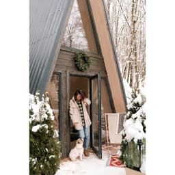 A woman stands outside an A-frame cabin, surrounded by snow, with a dog at her feet and evergreen decorations.