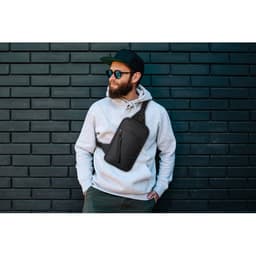 A man wears a grey hoodie with a black sling bag, standing against a dark brick wall. He has sunglasses and a cap.
