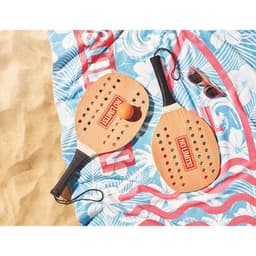 Two wooden beach paddles with a ball, resting on a patterned towel, alongside a pair of sunglasses.