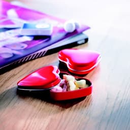 A heart-shaped tin in red, filled with assorted colourful sweets, rests on a wooden surface beside a magazine.