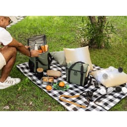 A thermal backpack and cooler bag in olive green, alongside picnic items on a checkered blanket in a grassy setting.