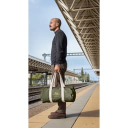 A man carries a green duffle bag with light grey straps at a train station. The bag features a logo.