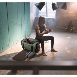 A green high-density 600D recycled polyester duffle bag sits on a fitness bench next to a woman preparing for a workout.