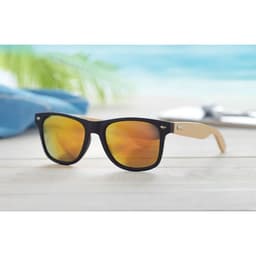 Sunglasses with black frames and brown arms, featuring yellow-tinted lenses. The background suggests a beach setting.