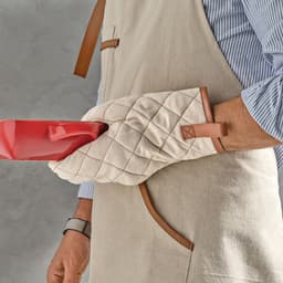 A beige apron with rPU details, featuring a person holding a red dish with quilted oven mitts.