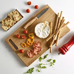 A wooden cutting board features a cheese wheel, cherry tomatoes, sliced ham, crispy breadsticks, and a knife, with a pepper mill nearby.