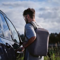 A man with glasses is holding a car key while wearing a grey backpack. The backpack features a TSA lock and anticut fabric.