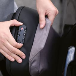 A black backpack with a TSA lock is being opened, revealing a grey fabric interior. The backpack has a branding logo.