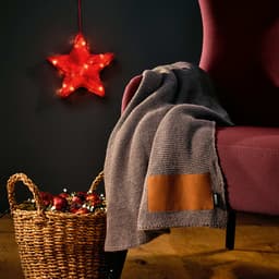 A grey blanket made from rPET and wool blend drapes over a burgundy chair, with a brown customisable patch.