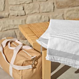 A cotton blanket in white and grey laid on a wooden table beside a tan bag with white straps.