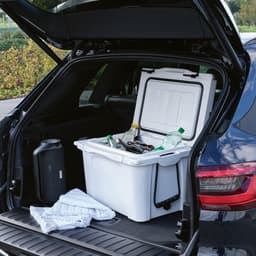 A white cooler sits in an open car boot, filled with various drinks and accompanied by towels. The car has a logo on it.